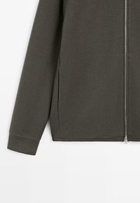 Giacca verde scuro con cerniera realizzata in tessuto morbido, con maniche lunghe, tasche laterali e una finitura liscia leggermente testurizzata.