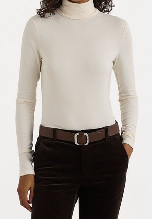 Femme portant un pull col roulé crème à manches longues, rentré dans un pantalon en velours côtelé marron foncé avec une ceinture marron à boucle argentée.