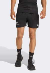 Zwarte sportieve shorts met een elastische tailleband, voorzien van witte logo-accenten. Gecombineerd met zwarte sokken en hardloopschoenen, wat een sportieve stijl benadrukt.