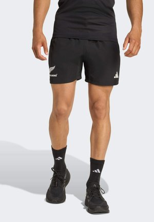 Shorts de sport noirs avec une taille élastique, présentant des accents de logo blanc. Portés avec des chaussettes noires et des chaussures de course, mettant en avant un style sportif.