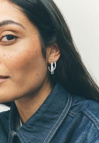 Orecchino a cerchio argento con design intrecciato, texture liscia e finitura lucida. Indossato su un orecchio con capelli scuri e colletto di camicia in denim visibile.