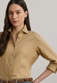 Bež srajca na gumbe iz mehkega, teksturiranega blaga. Ima ovratnik, rahlo zavihane dolge rokave in oprijet kroj.