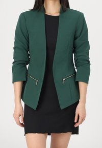 Zelený blazer se strukturovaným designem, dlouhými rukávy a kapsami na zip. V kombinaci s černými šaty, které zvýrazňují přesný střih.