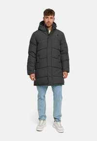 Manteau en duvet noir avec une capuche, présentant un design matelassé, une fermeture éclair sur le devant, des poches latérales et un logo sur la poitrine gauche.