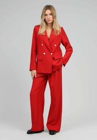 Femme avec de longs cheveux blonds portant un blazer rouge à double boutonnage et un pantalon large assorti, debout avec une main dans sa poche.