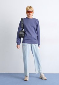 Paarse sweatshirt, lichtblauwe wijde broek, crème sneakers, kleine zwarte tas en oranje getinte zonnebril tegen een effen witte achtergrond.