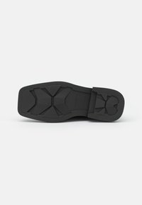 Semelle de chaussure noire avec surface en caoutchouc texturée présentant des motifs angulaires et un talon renforcé. Profil plat avec un design améliorant l'adhérence.