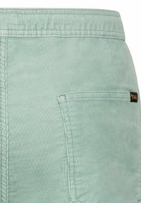 Close-up van lichtgroene ribfluwelen broek met zichtbare tailleband, achterzak en een zwart-oranje merklapje aan de rechterkant.