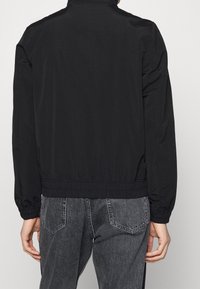 Osoba v černé lehké bundě s elastickými manžetami a pasem, v kombinaci se zesvětlenými černými džíny s zadními kapsami.