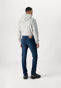 Homem a usar um capuz cinza, jeans justos azul-escuro com etiqueta de couro, e tênis pretos, de pé contra um fundo branco liso.