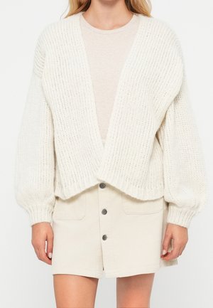 Kobieta w kremowym prążkowanym kardiganie z balonowymi rękawami, na beżowej bluzce i beżowej spódnicy sztruksowej z przodu zapinanej na guziki.