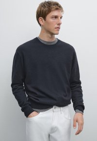 Maglione grigio scuro a girocollo realizzato in tessuto morbido, con maniche lunghe e polsini a costine. Indossato sopra una t-shirt grigia chiara e pantaloni bianchi.