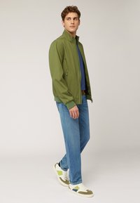 Giacca bomber verde con colletto rialzato, abbinata a una camicia blu e jeans azzurri chiari. Sneakers bianche con accenti verdi e gialli.