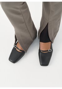 Chaussures en cuir noir à bout carré avec une fine bride à la cheville et un détail de boucle argentée. Associées à un pantalon gris avec une fente à l'ourlet.