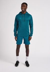 Felpa sportiva color teal con coulisse regolabile, abbinata a pantaloni corti coordinati. Presenta un tessuto morbido, cuciture piatte e un accento con logo.
