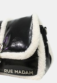 Musta patentoitu nahkalaukku valkoisella fleece-reunuksella, läppäkiinnitys, hopeiset yksityiskohdat ja "RUE MADAM" -logo etupuolella.