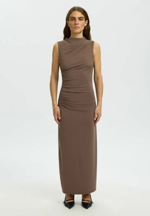 Frau mit langen braunen Haaren, die ein ärmelloses, bodenlanges braunes Kleid und schwarze spitze Schuhe trägt, steht vor einem schlichten hellen Hintergrund.