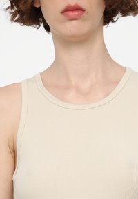 Gros plan d'une personne aux cheveux courts brun portant un haut côtelé sans manches beige, montrant le cou, les épaules et les lèvres.