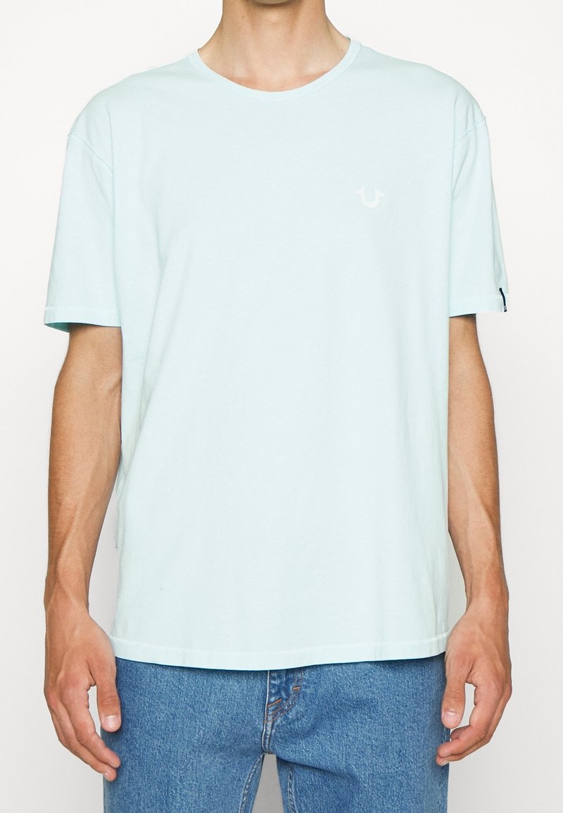 Světlemodré bavlněné tričko s kulatým výstřihem, krátkými rukávy a malým bílým logem na hrudi. Volný střih, měkká textura.