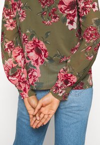 Blusa floreale con uno sfondo verde e grandi fiori rosa e rossi, maniche lunghe con polsini arricciati, abbinata a jeans blu.