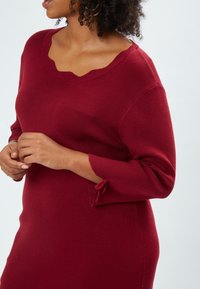 Frau in einem figurbetonten roten Kleid mit gewelltem Ausschnitt und kleiner Schleifenapplikation am Ärmel, die vor einem schlichten hellen Hintergrund steht.