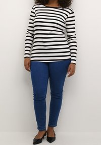 Chemise à manches longues rayée noir et blanc associée à un jean skinny bleu et des ballerines noires à bouts pointus, sur un fond blanc uni.