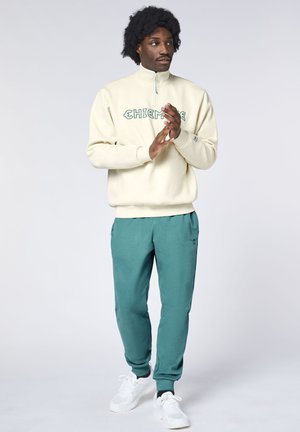 Man standing wearing cream half-zip sweatshirt, teal joggers, and white sneakers, with hands clasped and looking to the side.
