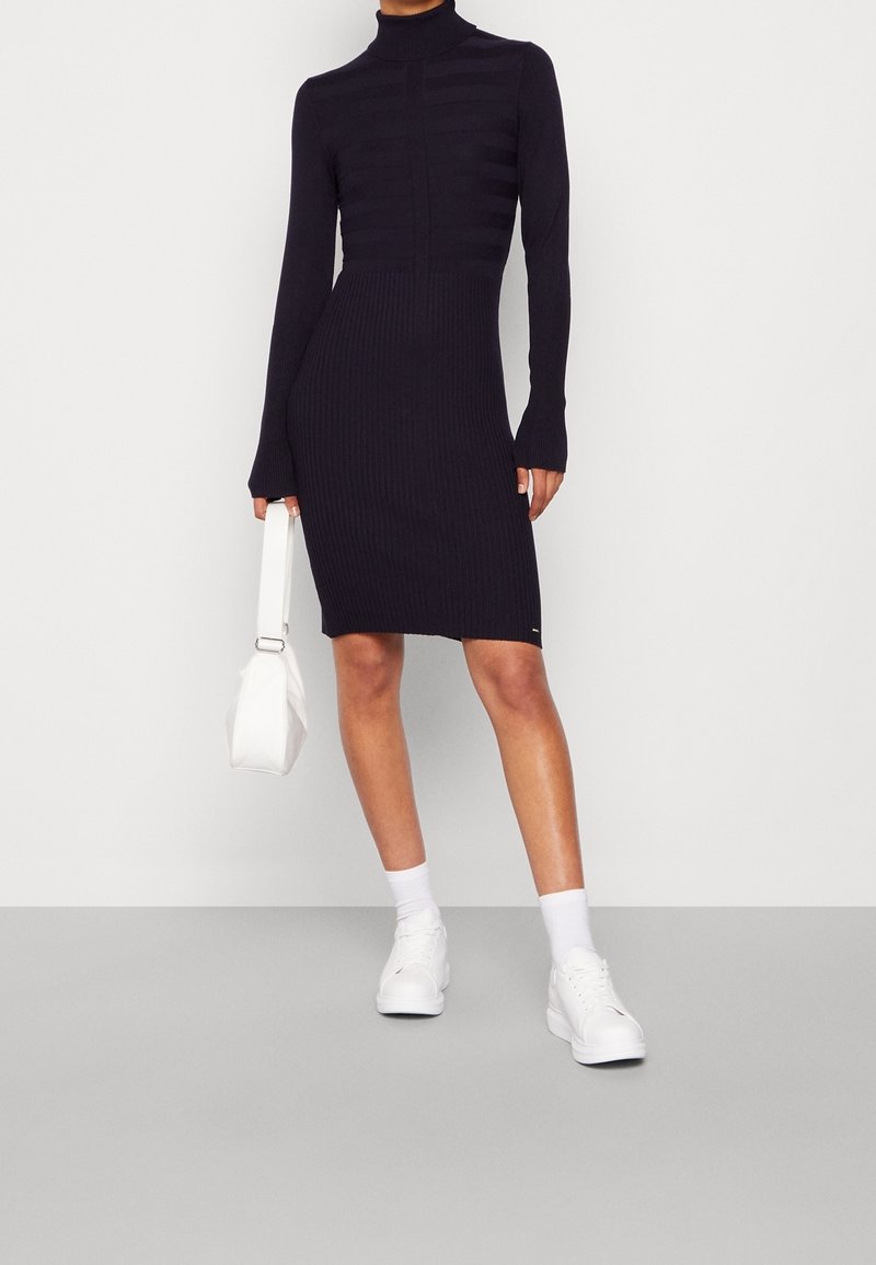 Femme portant une robe noire côtelée ajustée à col roulé avec manches longues, tenant un sac à main blanc, et chaussée de baskets et de chaussettes blanches.