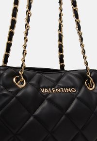 Mustahuoneilla nahkainen käsilaukku kultaketjuisilla hihnoilla ja logoyksityiskohdalla. Varustettu teksturoidulla pinnalla ja pyöreillä metalliosilla.