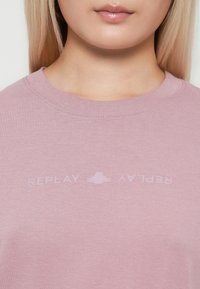 Tuvo plāna attēls ar personu, kas valkā gaiši violetu T-kreklu ar smalku, apgrieztu "REPLAY" tekstu un logotipu uz krūtīm.