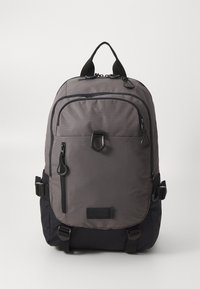 Sac à dos gris avec une surface texturée, plusieurs poches zippées, des sangles ajustables et un petit accent noir en bas.