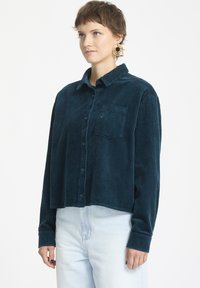Teal kordtøy skjorte med knapper och krage, enkel bröstficka och långa ärmar, i kombination med ljusblå jeans. Texturerat ribbat tyg.
