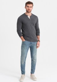 Grafiittivärinen pitkähihainen henley-paita valkoisella nappilistalla, yhdistettynä vaaleansinisiin slim-fit-farkkuihin ja vaaleisiin lenkkareihin. Yksinkertainen tausta.