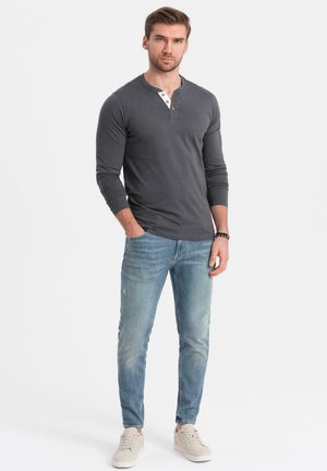 Camiseta de manga larga tipo henley en grafito con tapeta de botones blancos, combinada con jeans ajustados de color azul claro y zapatillas beige. Fondo liso.