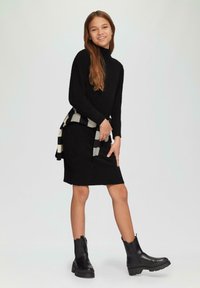 Zwarte gebreide jurk met een geribbelde zoom en een hoge hals, gecombineerd met een zwart-wit gestreepte sjaal en stevige zwarte enkellaarzen.