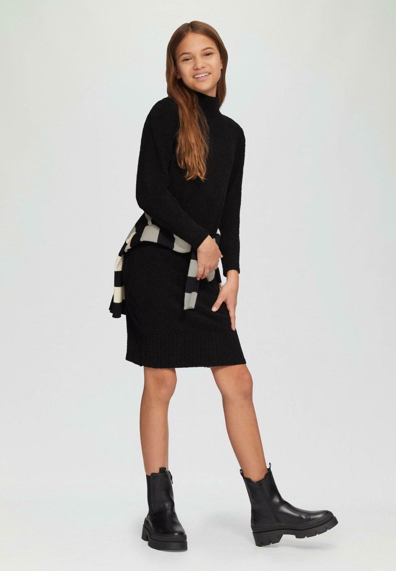 Zwarte gebreide jurk met een geribbelde zoom en een hoge hals, gecombineerd met een zwart-wit gestreepte sjaal en stevige zwarte enkellaarzen.
