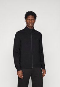 Jacket preta com fecho éclair e gola alta, tecido texturizado e bolsos laterais, apresentando uma marcação discreta no peito e punhos ajustados.