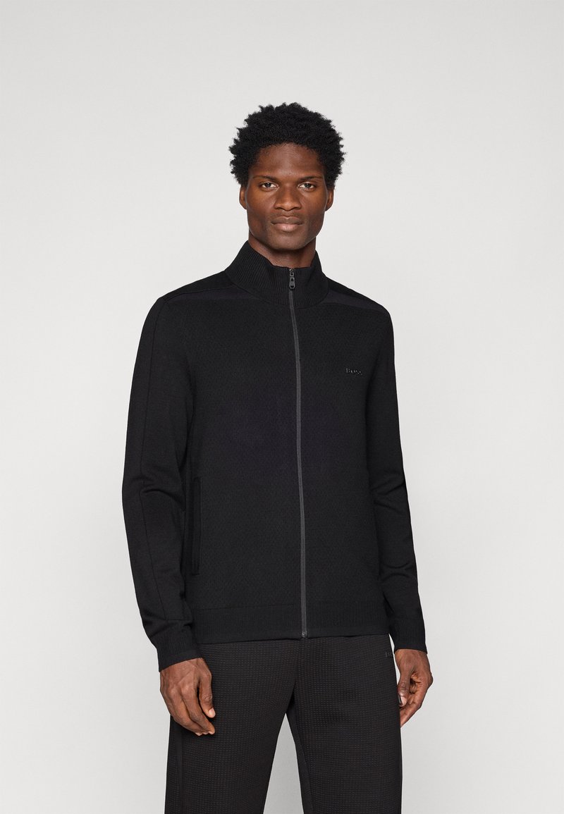 Jacket preta com fecho éclair e gola alta, tecido texturizado e bolsos laterais, apresentando uma marcação discreta no peito e punhos ajustados.