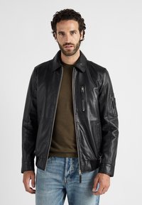 Schwarze Lederjacke mit Reißverschluss vorne, spitzem Kragen und gerippten Bündchen. Verfügt über Seitentaschen und eine Reißverschlusstasche am Oberarm.
