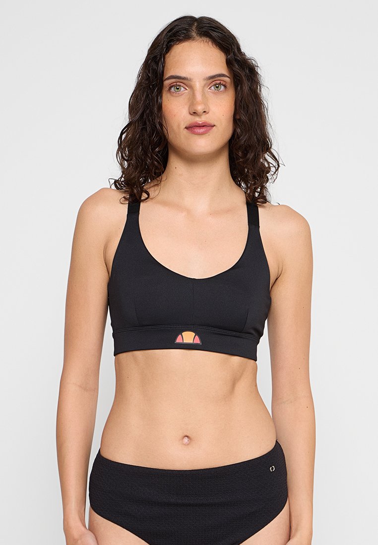 Kobieta z kręconymi ciemnymi włosami, ubrana w czarny stanik sportowy i czarne teksturowane spodenki z wysokim stanem na jednolitym tle.