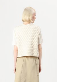 Pute beige vest med et teksturert diamantmønster, båret over en stripete t-skjorte med korte ermer, sammen med et khaki skjørt.