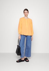 Blusa laranja de botão com mangas longas, combinada com jeans de cintura alta em azul denim de perna larga, sandálias pretas de slide e uma mala tote preta.