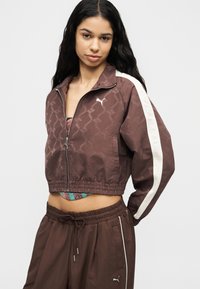 Νεαρή γυναίκα που φοράει καφέ φόρμα Puma με λευκές ρίγες στα μανίκια και το παντελόνι, cropped μπουφάν με φερμουάρ στη μέση, στέκεται μπροστά σε απλό φόντο.