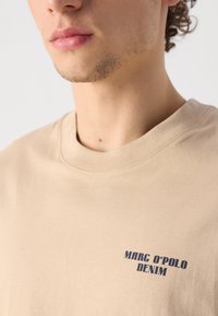 Beige bomulds t-shirt med rund halsudskæring og korte ærmer; har mørkeblå tekst "MARC O'POLO DENIM" på forsiden.