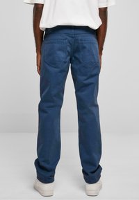 Pantalons en coton bleu avec une coupe droite, comportant deux grandes poches plaquées à l'arrière et une ceinture avec passants. Des chaussures blanches visibles.