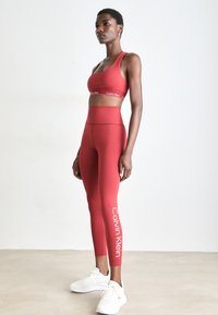 Vrouw staat in een rode Calvin Klein sport-bh en leggings met witte sneakers tegen een effen witte achtergrond.