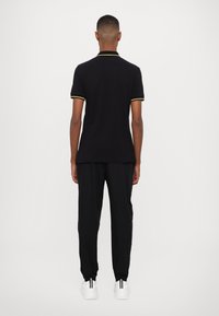 Tricou polo negru cu margini galbene pe mâneci, asortat cu pantaloni de trening negri și adidași albi cu dungi negre, văzut din spate.