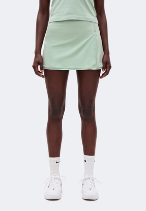 Personne portant une jupe de sport vert clair, un haut assorti, des chaussettes blanches Nike et des baskets blanches, debout devant un fond uni.