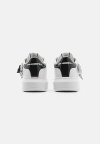 Adidași albi cu accente din piele patent neagră, având o textură netedă, formă rotundă și toc cu marcaj. Detaliu notabil cu fermoar.
