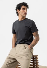 Donkergrijze t-shirt met een klassieke ronde hals, gecombineerd met beige broek. Glad materiaal en een getailleerd ontwerp, met zichtbare armtattoos.